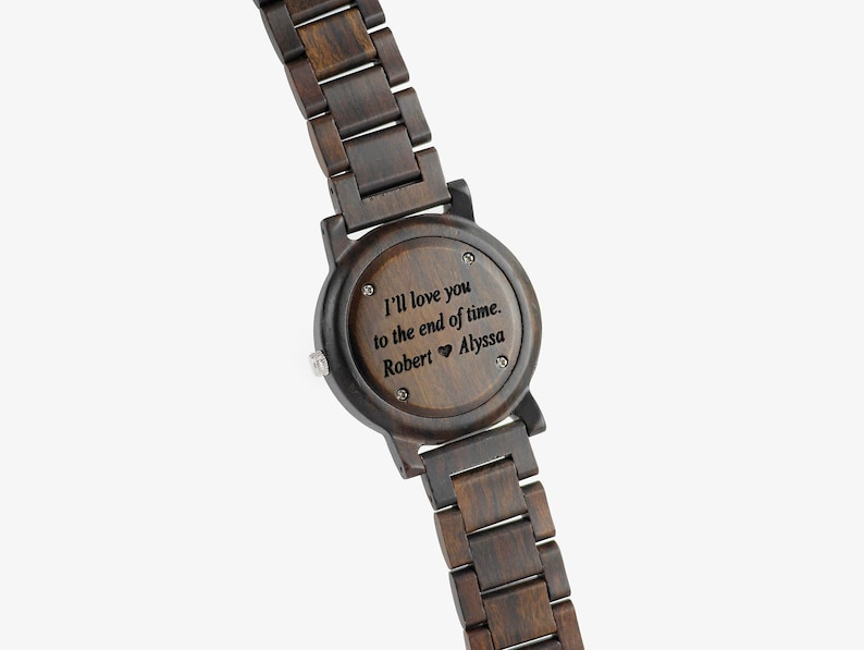 Puede incluir: Un reloj de pulsera de madera marr&oacute;n oscuro con una esfera redonda y un mensaje grabado en negro que dice "Te amar&eacute; hasta el final de los tiempos. Robert &hearts; Alyssa".