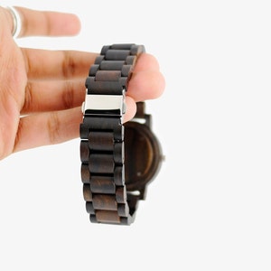 Puede incluir: Un reloj de pulsera de madera marr&oacute;n oscuro con una hebilla plateada. El reloj tiene una esfera redonda y una correa hecha de eslabones de madera conectados.