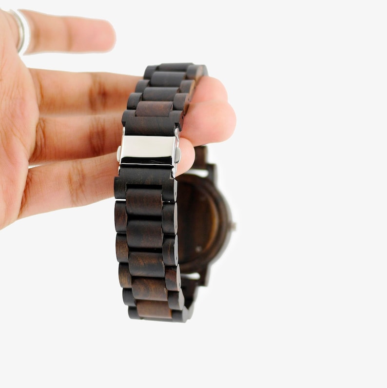 Puede incluir: Un reloj de pulsera de madera marr&oacute;n oscuro con una hebilla plateada. El reloj tiene una esfera redonda y una correa hecha de eslabones de madera conectados.