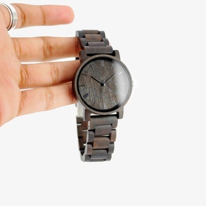 Puede incluir: Un reloj de pulsera de madera marr&oacute;n oscuro con una esfera negra y marcadores de hora en n&uacute;meros romanos. El reloj tiene una correa de madera.