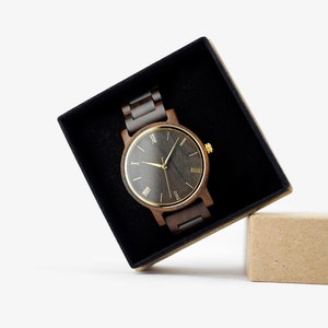 Puede incluir: Un reloj de pulsera de madera oscura con una esfera negra y marcadores de hora en n&uacute;meros romanos dorados. El reloj est&aacute; en una caja de terciopelo negro con una caja de cart&oacute;n marr&oacute;n a su lado.
