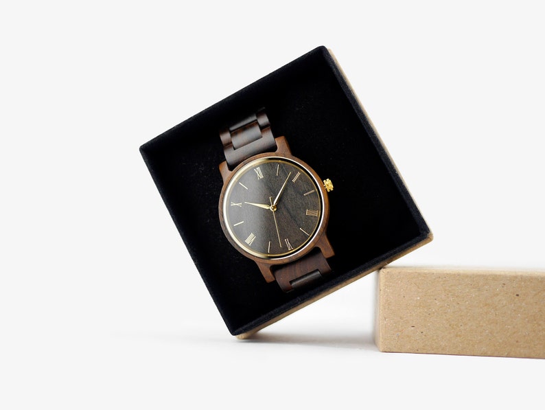 Puede incluir: Un reloj de pulsera de madera oscura con una esfera negra y marcadores de hora en n&uacute;meros romanos dorados. El reloj est&aacute; en una caja de terciopelo negro con una caja de cart&oacute;n marr&oacute;n a su lado.
