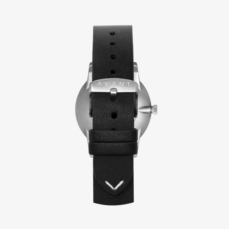 May include: A silver watch with a black leather band. The watch face is plain and the band has a white stitching detail. The watch has the word "AVANT" printed on the back.