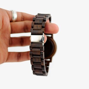 Puede incluir: Un reloj de pulsera de madera marr&oacute;n oscuro con un cierre plateado. La correa del reloj est&aacute; hecha de eslabones de madera individuales.
