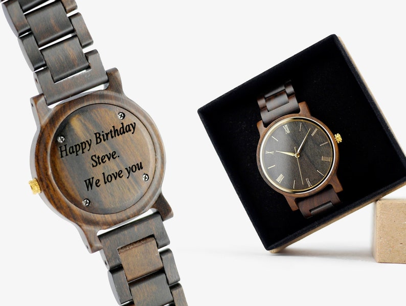 Puede incluir: Un reloj de pulsera de madera marr&oacute;n oscuro con una esfera negra y marcadores de hora en n&uacute;meros romanos dorados. El reloj est&aacute; grabado con el mensaje "Happy Birthday Steve. We love you".
