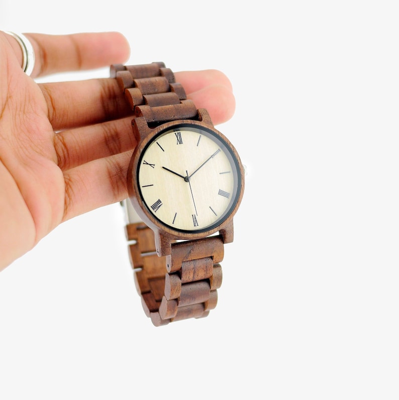 Puede incluir: Un reloj de pulsera de madera marr&oacute;n con una esfera de color crema y marcadores de hora romanos negros.