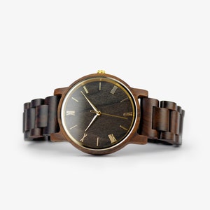 Puede incluir: Un reloj de pulsera de madera marr&oacute;n con una esfera negra y marcadores de hora en n&uacute;meros romanos dorados.