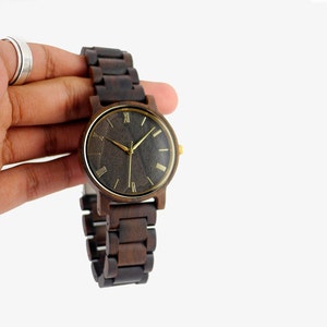 Puede incluir: Un reloj de pulsera de madera marr&oacute;n oscuro con una esfera negra y marcadores de hora en n&uacute;meros romanos dorados. El reloj tiene una correa de madera marr&oacute;n.