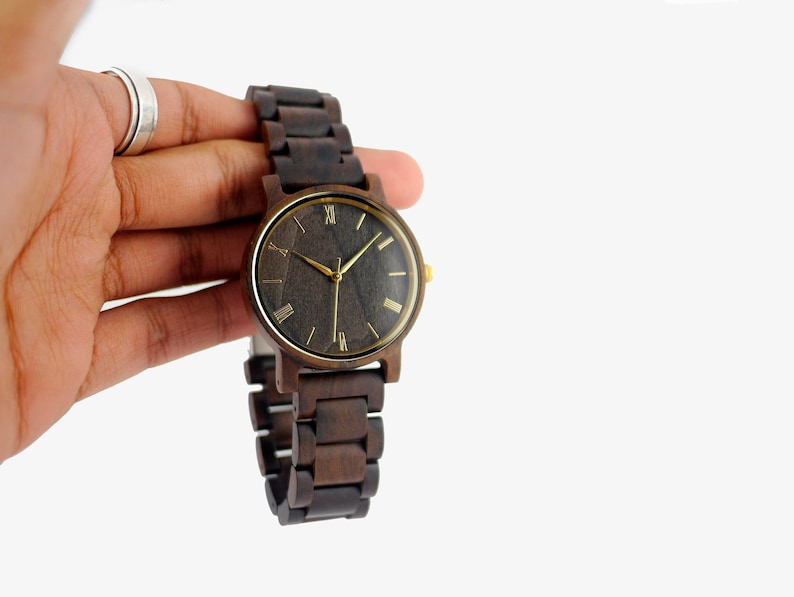 Puede incluir: Un reloj de pulsera de madera marr&oacute;n oscuro con una esfera negra y marcadores de hora en n&uacute;meros romanos dorados. El reloj tiene una correa de madera marr&oacute;n.