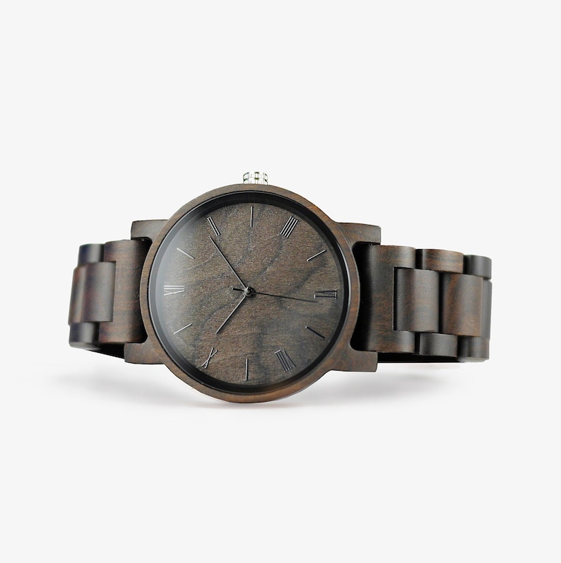 Puede incluir: Un reloj de pulsera de madera marr&oacute;n oscuro con marcadores de hora en n&uacute;meros romanos y una esfera negra.