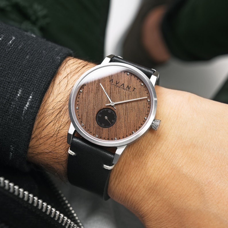 May include: A silver wristwatch with a black leather band. The watch face is made of wood with a silver trim and the word "AVANT" printed on it.