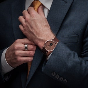 May include: A rose gold watch with a wooden face and brown leather strap. The watch is on a wrist next to a silver ring. The person is wearing a navy blue suit and a yellow tie.