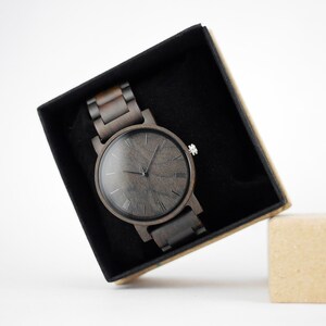 Puede incluir: Un reloj de pulsera de madera negro con n&uacute;meros romanos en la esfera, en una caja negra con una caja de cart&oacute;n marr&oacute;n al lado.