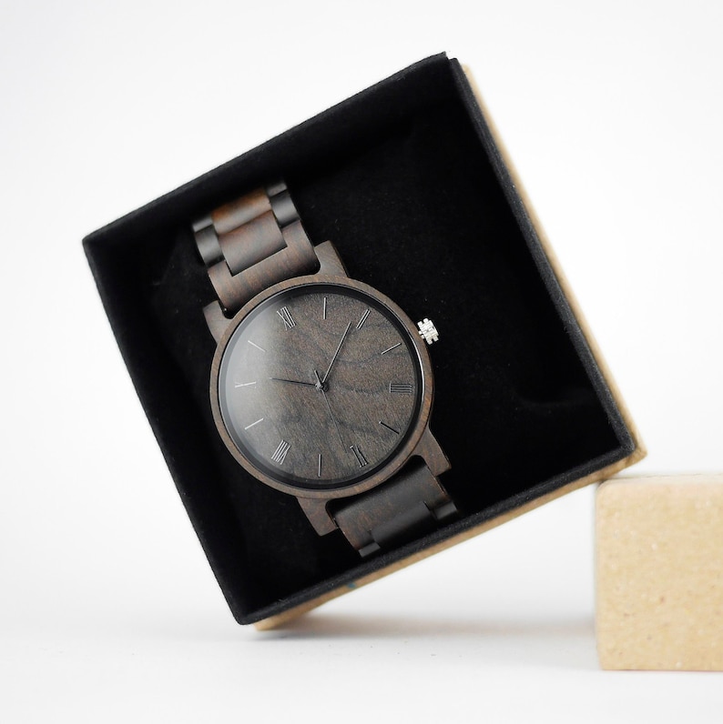 Puede incluir: Un reloj de pulsera de madera negro con n&uacute;meros romanos en la esfera, en una caja negra con una caja de cart&oacute;n marr&oacute;n al lado.