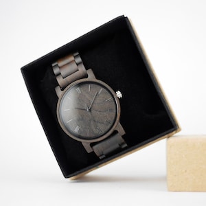 Puede incluir: Un reloj de pulsera de madera negro con n&uacute;meros romanos en la esfera, en una caja de terciopelo negro.