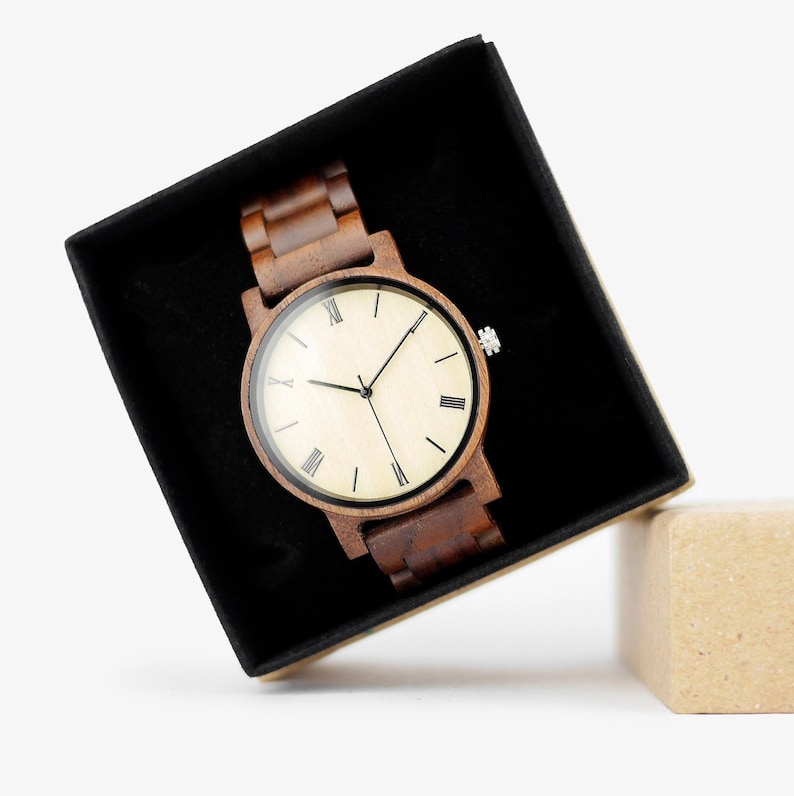 Puede incluir: Un reloj de pulsera de madera marr&oacute;n con una esfera blanca y marcadores de hora en n&uacute;meros romanos negros. El reloj est&aacute; en una caja negra con una caja de cart&oacute;n marr&oacute;n al lado.