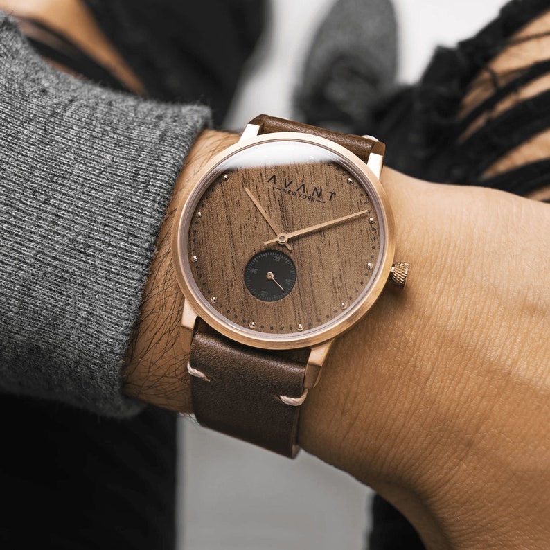 May include: A rose gold wristwatch with a wooden face and a brown leather strap. The watch face features the text "AVANT NEW YORK" and a small subdial. The watch is on a wrist, with a gray sweater in the background.