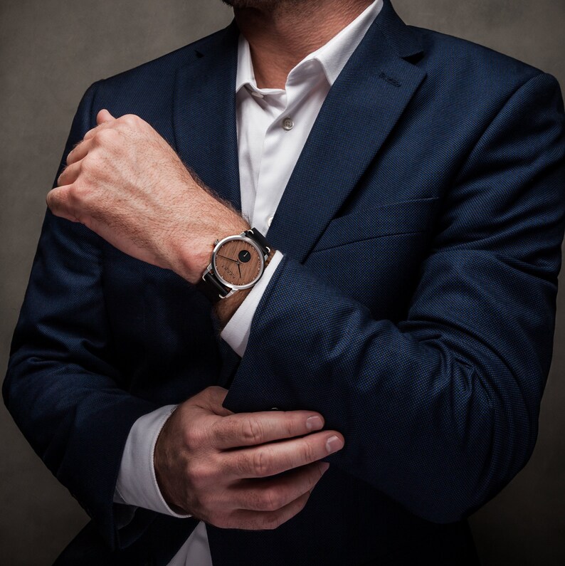 May include: A man wearing a dark blue suit jacket with a white dress shirt and a wristwatch with a wooden face and black leather band.