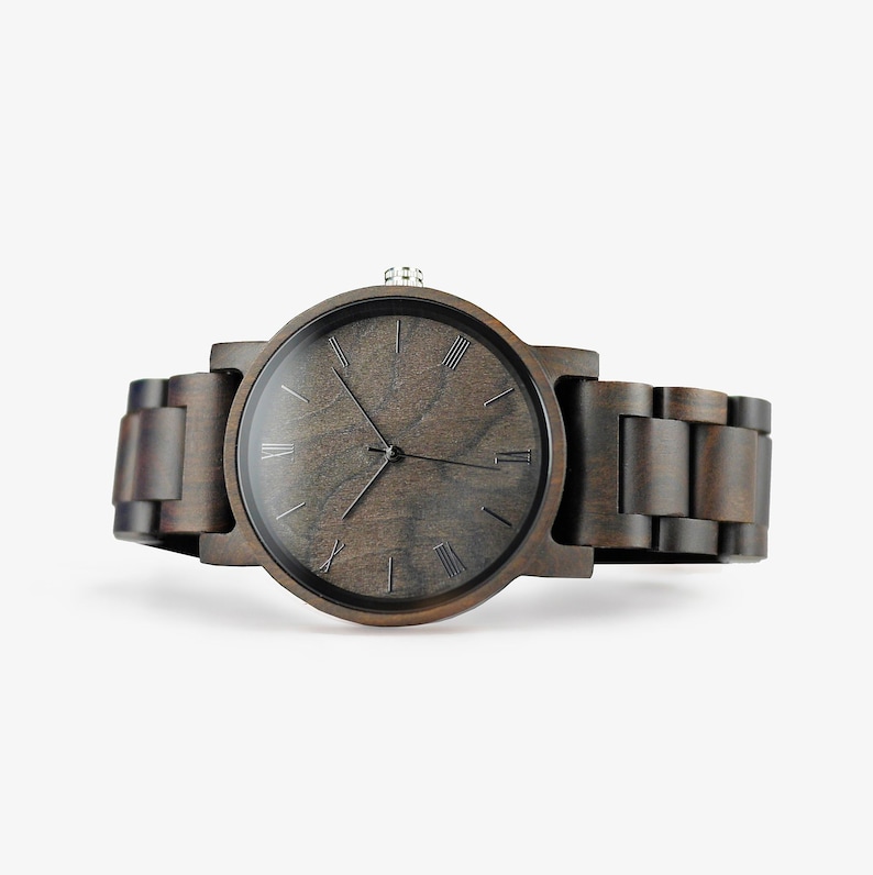 Puede incluir: Un reloj de pulsera de madera marr&oacute;n oscuro con una esfera redonda y marcadores de hora en n&uacute;meros romanos. El reloj tiene una correa de madera marr&oacute;n oscuro.