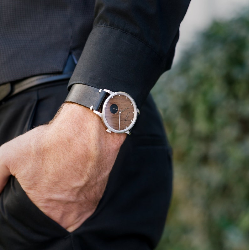 May include: A silver wristwatch with a wooden face and black leather band. The watch face has a black circle in the center and a silver hand.