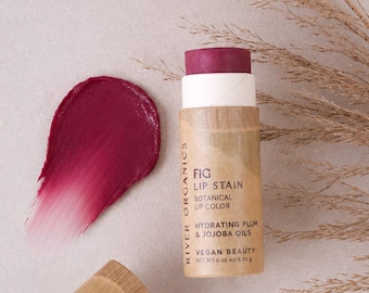 May include: A cylindrical River Organics Fig Lip Stain tube with a tan paper label, a plum-colored lip product, and a matching cap. The label reads Fig Lip Stain, Botanical Lip Color, Hydrating Plum & Jojoba Oils, Vegan Beauty, Net Wt 0.30 oz.