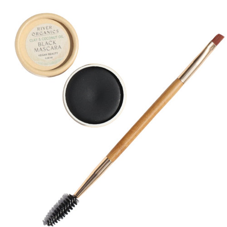 Puede incluir: Un peque&ntilde;o recipiente redondo de m&aacute;scara de pesta&ntilde;as negra con un cepillo aplicador de madera. El recipiente est&aacute; etiquetado como "River Organics Clay & Coconut Oil Black Mascara Vegan Beauty".