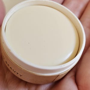 May include: A round, off-white beauty product in a small, open container. The product has a smooth surface and is surrounded by a white rim and a light brown cardboard-like container. The word "BEAUTY" is partially visible on the container.