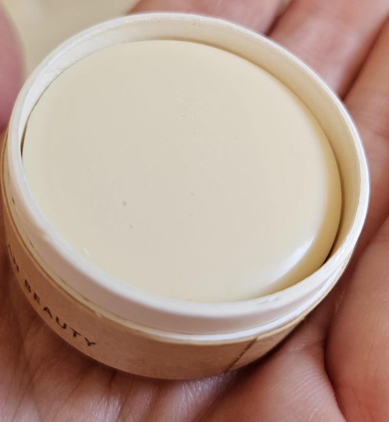 May include: A round, off-white beauty product in a small, open container. The product has a smooth surface and is surrounded by a white rim and a light brown cardboard-like container. The word "BEAUTY" is partially visible on the container.