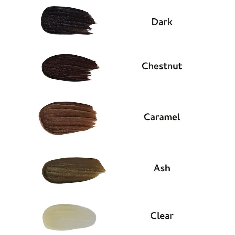 May include: A white background displays five swatches of eyebrow gel in various shades. The colors are dark, chestnut, caramel, ash, and clear. Each swatch is accompanied by its corresponding color name in black text.