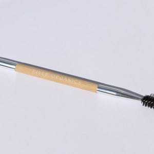 May include: A double-ended makeup tool with a silver handle and a natural bamboo center. One end features a small, angled brush, and the other has a black, bristled spoolie. The text "RIVER ORGANICS" is printed on the bamboo.