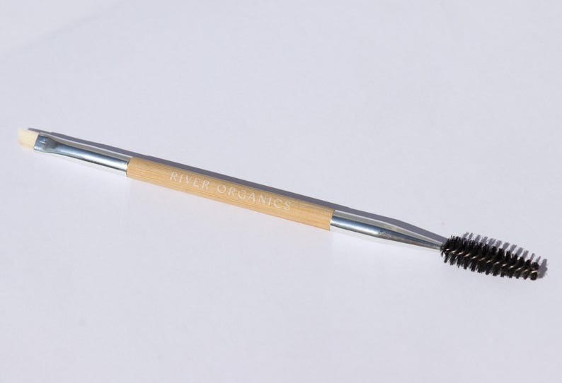 May include: A double-ended makeup tool with a silver handle and a natural bamboo center. One end features a small, angled brush, and the other has a black, bristled spoolie. The text "RIVER ORGANICS" is printed on the bamboo.