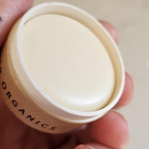 May include: A close-up of a small, round container of cream-colored balm. The container is made of cardboard with the word "ORGANICS" printed on the side. The balm is smooth and appears to be solid.