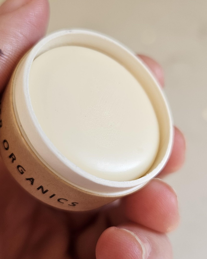 May include: A close-up of a small, round container of cream-colored balm. The container is made of cardboard with the word "ORGANICS" printed on the side. The balm is smooth and appears to be solid.