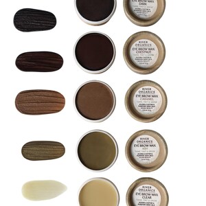 May include: An assortment of River Organics eyebrow waxes in various shades, including Dark, Chestnut, Caramel, Ash, and Clear. Each wax is in a small, round container with a natural paper label. Swatches of each color are displayed next to the containers.
