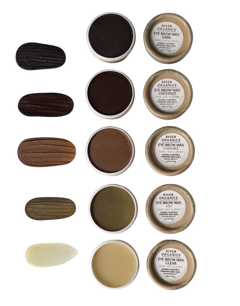 May include: An assortment of River Organics eyebrow waxes in various shades, including Dark, Chestnut, Caramel, Ash, and Clear. Each wax is in a small, round container with a natural paper label. Swatches of each color are displayed next to the containers.