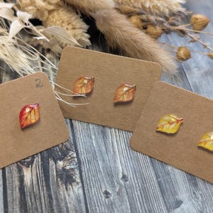 Puede incluir: Tres pares de pendientes de bot&oacute;n con forma de hoja en tonos rojos, naranjas y amarillos, expuestos en tarjetas marrones. Los pendientes tienen un acabado brillante y est&aacute;n sobre un fondo de madera r&uacute;stica, con flores secas.