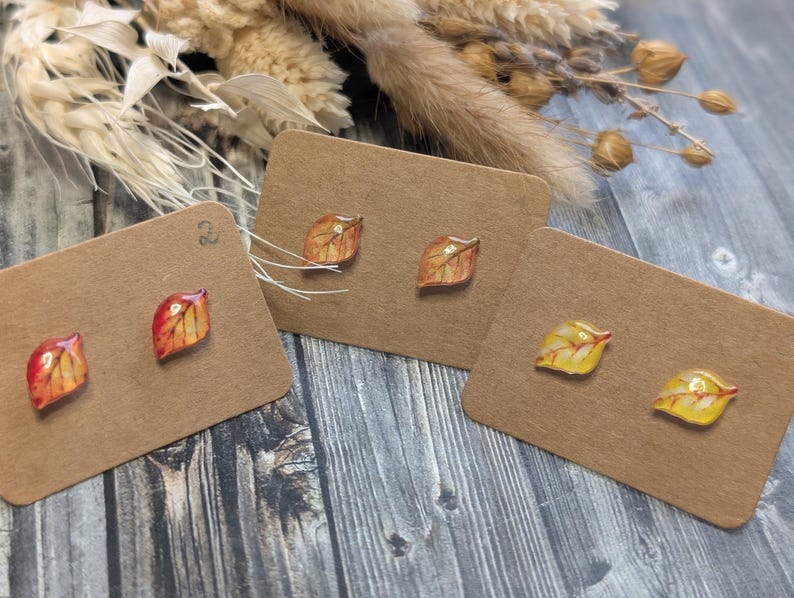 Puede incluir: Tres pares de pendientes de bot&oacute;n con forma de hoja en tonos rojos, naranjas y amarillos, expuestos en tarjetas marrones. Los pendientes tienen un acabado brillante y est&aacute;n sobre un fondo de madera r&uacute;stica, con flores secas.