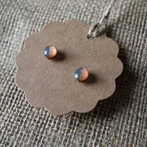 May include: A pair of small, round stud earrings with a copper and gray color scheme. The earrings are displayed on a brown paper tag.