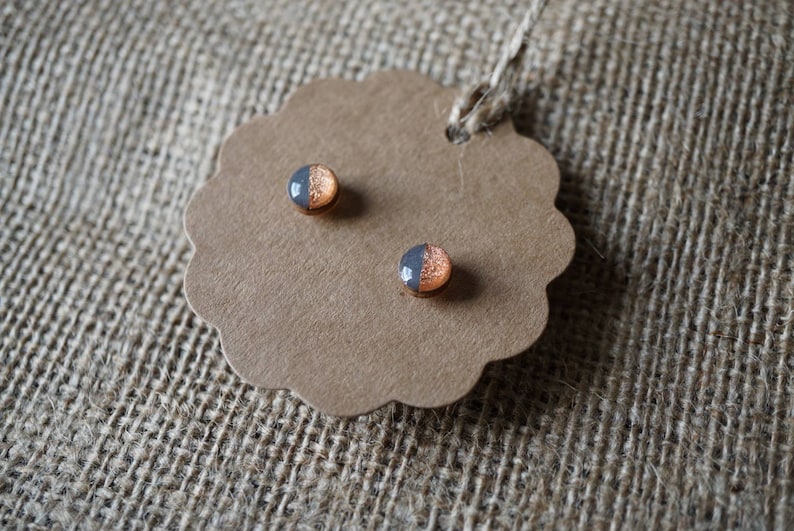 May include: A pair of small, round stud earrings with a copper and gray color scheme. The earrings are displayed on a brown paper tag.
