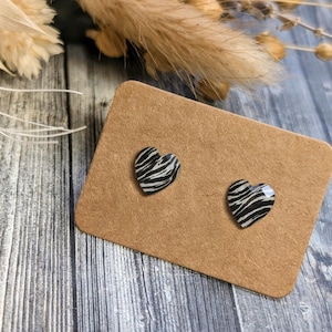 Könnte beinhalten: Ohrstecker in Herzform mit Zebra-Print-Design in Schwarz und Weiß. Sie liegen auf einer braunen Karte. Der Hintergrund ist eine Holzoberfläche mit getrockneten Pflanzen.