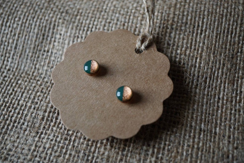 May include: A pair of small, round earrings with a green and gold design. The earrings are on a brown card with a scalloped edge.
