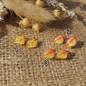 Puede incluir: Tres pares de pendientes en forma de hoja. El primer par es amarillo y dorado, el segundo par es rojo y naranja, y el tercer par es amarillo y naranja.