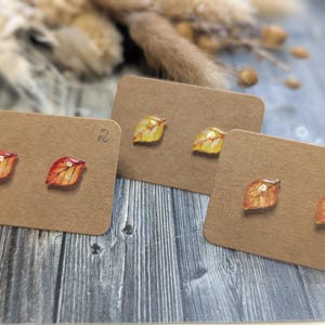Puede incluir: Tres pares de pendientes de bot&oacute;n con forma de hoja expuestos en tarjetas marrones. Los pendientes presentan colores oto&ntilde;ales, incluyendo rojo, naranja y amarillo. El fondo incluye plantas secas y una superficie de madera.