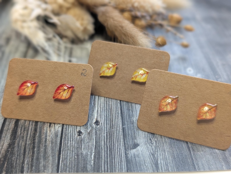 Puede incluir: Tres pares de pendientes de bot&oacute;n con forma de hoja expuestos en tarjetas marrones. Los pendientes presentan colores oto&ntilde;ales, incluyendo rojo, naranja y amarillo. El fondo incluye plantas secas y una superficie de madera.