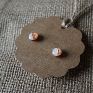 May include: A pair of small, round earrings with a white and copper design. The earrings are displayed on a brown card.