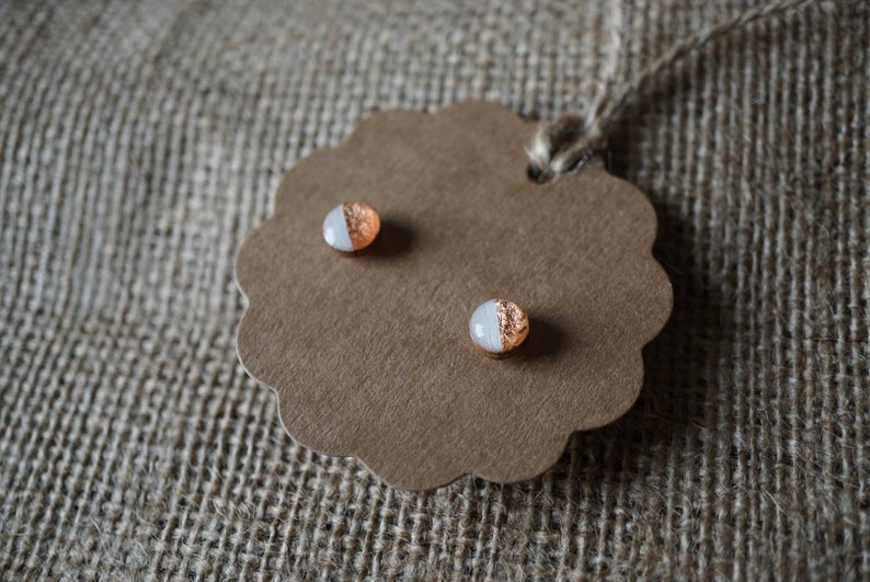 May include: A pair of small, round earrings with a white and copper design. The earrings are displayed on a brown card.