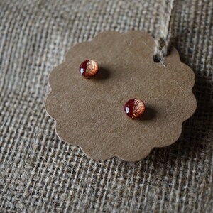 May include: A pair of small, round earrings with a red and copper metallic finish. The earrings are displayed on a brown paper tag with a scalloped edge.