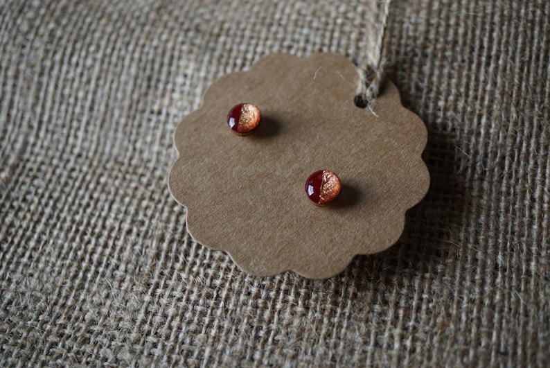 May include: A pair of small, round earrings with a red and copper metallic finish. The earrings are displayed on a brown paper tag with a scalloped edge.
