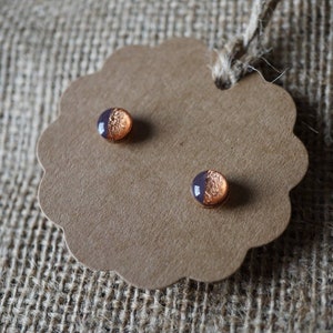 May include: A pair of small, round earrings with a copper and purple design. The earrings are displayed on a brown card.