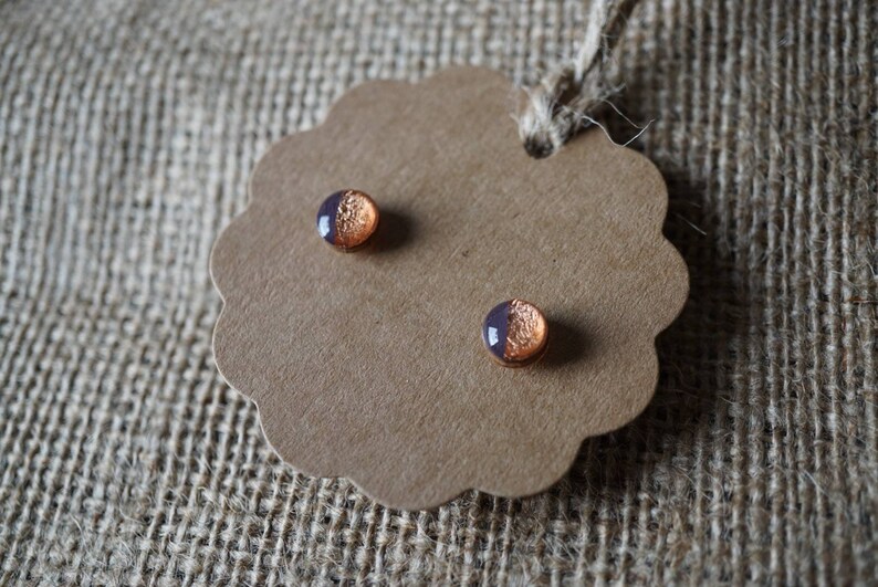 May include: A pair of small, round earrings with a copper and purple design. The earrings are displayed on a brown card.